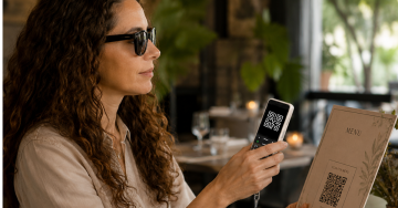Eine blinde Frau mit Sonnenbrille sitzt in einem Restaurant und scannt mit ihrem BlindShell-Telefon einen QR-Code auf einer Speisekarte. Auf dem Telefonbildschirm ist zu sehen, wie ein QR-Code erkannt wird. Das Bild zeigt, wie der NFC- und QR-Lesegerät auf BlindShell genutzt werden kann, um auf Speisekarten und andere öffentliche Informationen zuzugreifen.