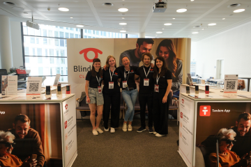 Das BlindShell-Team steht gemeinsam am BlindShell-Stand während der SightCity 2026 in Frankfurt. Fünf Teammitglieder mit Veranstaltungsausweisen lächeln und posieren vor einer großen BlindShell Classic 3-Kulisse. Der Stand bietet Produktpräsentationen, QR-Codes und Informationen zu BlindShell-Funktionen wie Tandem und Observo. Der helle Ausstellungsbereich ist bereit, Besucher zu empfangen, die sich für barrierefreie Technologie für blinde und sehbehinderte Nutzer interessieren.