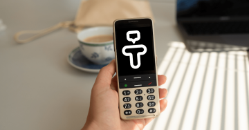 Eine Person hält ein BlindShell-Telefon in der Hand. Auf dem Display des Telefons wird ein Symbol für die Tandem-App angezeigt, das wie ein Buchstabe T mit einem Kopf aussieht.
