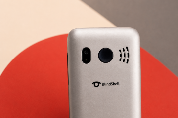 Nahaufnahme des oberen hinteren Bereichs eines silbernen BlindShell Classic Lite-Handys vor einem geometrischen Hintergrund in warmem Beige, Rot und Dunkelgrau. Das Bild hebt die runde SOS-Taste und das BlindShell-Logo hervor.