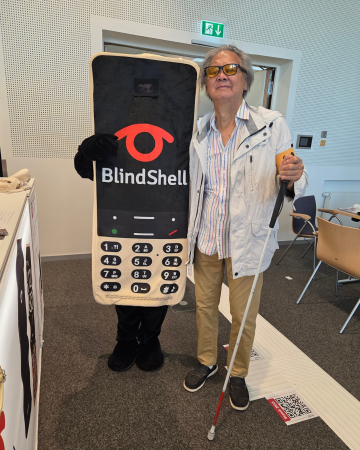 Einer unserer glücklichen BlindShell Classic 3-Gewinner posiert stolz neben dem neuen BlindShell-Maskottchen. Mit Sonnenbrille, einer leichten Jacke und einem weißen Stock in der Hand steht der Gewinner lächelnd neben dem Maskottchen. Zusammen fangen sie einen herzerwärmenden SightCity 2026-Moment ein, voller Aufregung, Dankbarkeit und dem Gemeinschaftsgefühl, das diese Veranstaltungen so besonders macht.