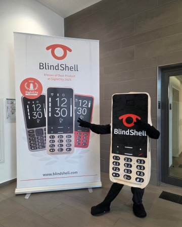 Unser neues BlindShell-Maskottchen, gestaltet als riesiges goldenes BlindShell-Handy mit Tastatur, Bildschirm und leuchtendem BlindShell-Logo, posiert stolz neben einem BlindShell-Banner. Mit seiner verspielten Haltung und seiner freundlichen Ausstrahlung wurde das Maskottchen schnell zum Liebling der Besucher, verbreitete gute Laune und sorgte für zusätzlichen Spaß auf der SightCity 2026.