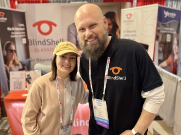 Molly Burke und Sam, the Blind Life, stehen eng beieinander und lächeln am BlindShell-Stand während einer Konferenz für assistive Technologien. Molly trägt eine helle Mütze und einen beigen Pullover und hat ein Konferenz-Namensschild an einem Umhängeband um den Hals. Sam trägt ein schwarzes BlindShell-Poloshirt mit dem BlindShell-Logo und ein Aussteller-Namensschild. Beide wirken entspannt und freundlich. Hinter ihnen sind BlindShell-Banner und Werbematerialien zu sehen.