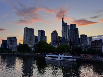 Frankfurts Skyline bei Sonnenuntergang, mit modernen Wolkenkratzern, die das letzte Licht des Tages reflektieren. Die markanten Hochhäuser der Stadt ragen über den Main, wo Boote am Ufer vor Anker liegen. Rosa und orangefarbene Wolken ziehen sich über den Abendhimmel und schaffen einen malerischen Anblick auf Deutschlands Finanzhauptstadt, die oft als „Mainhattan“ bezeichnet wird.