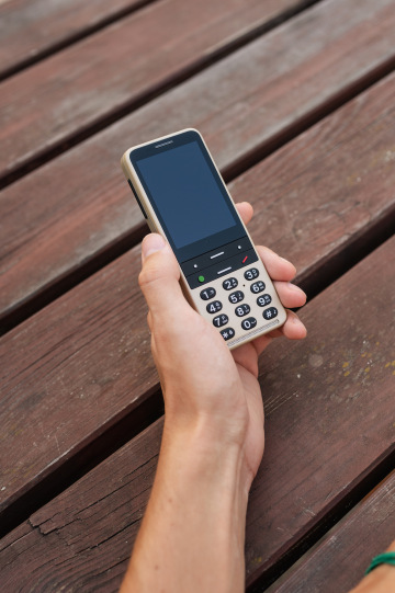 Eine Hand hält ein BlindShell Classic 3 Telefon in der Farbe „Sandgold“. Das Telefon hat große, taktile Tasten mit gut lesbaren Zahlen und Symbolen, ideal für sehbehinderte Benutzer. Der Hintergrund zeigt eine hölzerne Oberfläche.