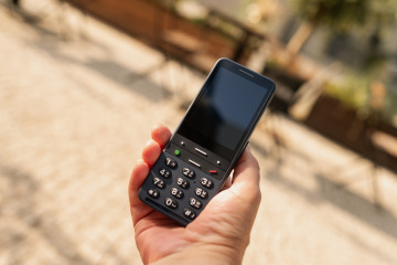 Eine Hand hält ein BlindShell Classic 3 Telefon in der Farbe „Ozeanblau“. Das Telefon hat große, taktile Tasten mit klar erkennbaren Zahlen und Symbolen, speziell entworfen für sehbehinderte Benutzer. Im Hintergrund sind unscharfe Tische und Stühle im Freien zu sehen.